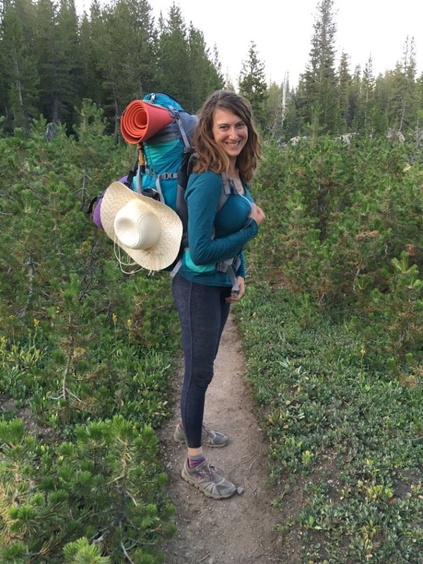A smiling woman wearing a hiking backpack on a narrow dirt path surrounded by foliage.