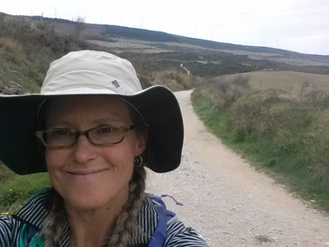 A selfie image of a woman wearing a fisherman's hat with a dirt trail and green fields behind her.