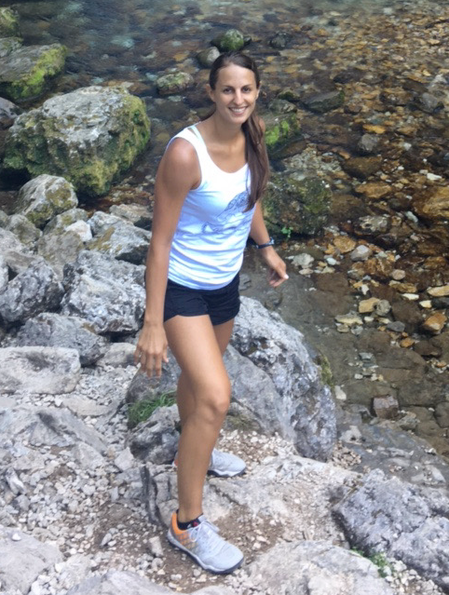 A woman wearing shorts and a tank top stands on a rocky terrain with a stream running behind her.
